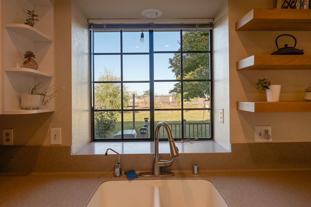Kitchen window with a view of a backyard, sink with a faucet, and shelving with decorative items.