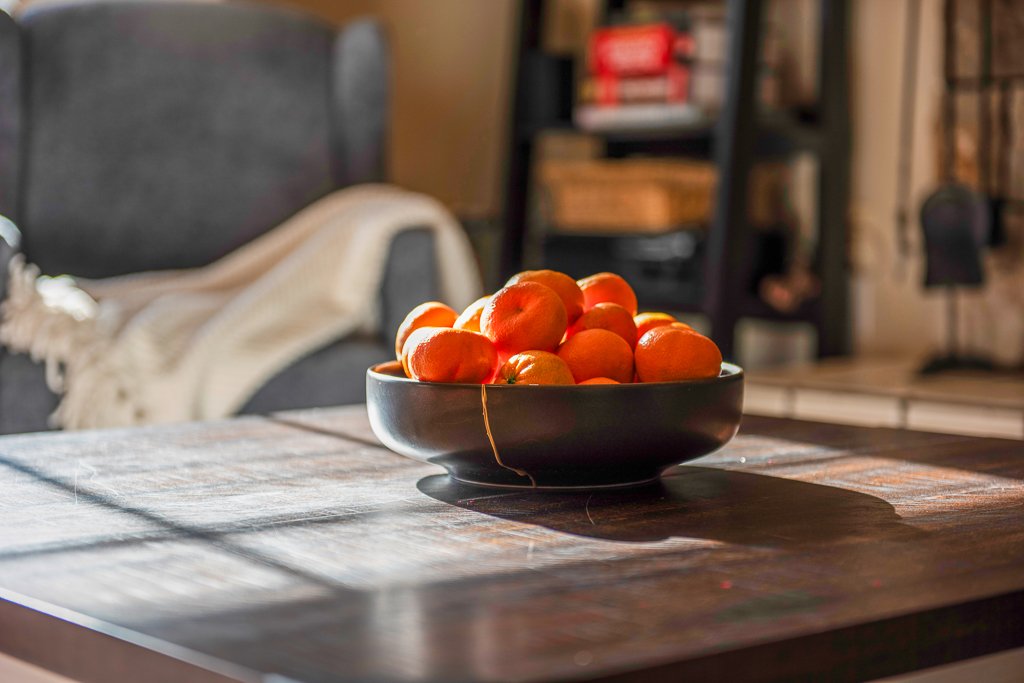 A black bowl filled with bright orange persimmons placed on a wooden table, with sunlight casting shadows.
