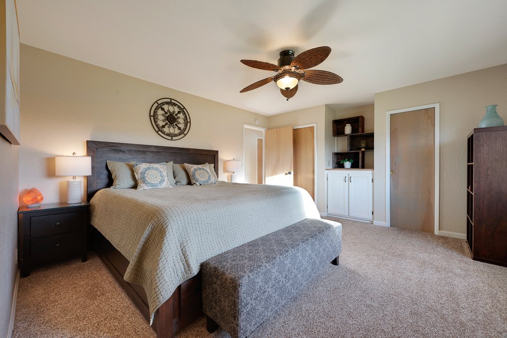 A bedroom with a large bed, dark wooden headboard and side tables, decorative pillows, a ceiling fan, and built-in shelving with cabinets.
