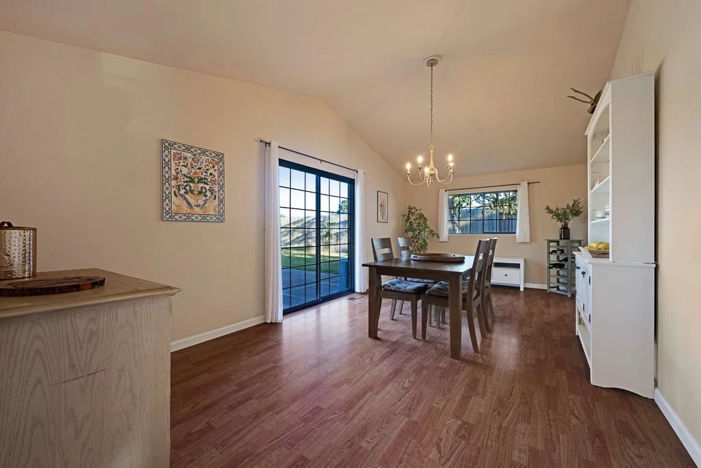 Dining room with wooden table, four chairs, chandelier, sliding glass door, window, white cabinet, and potted plants.