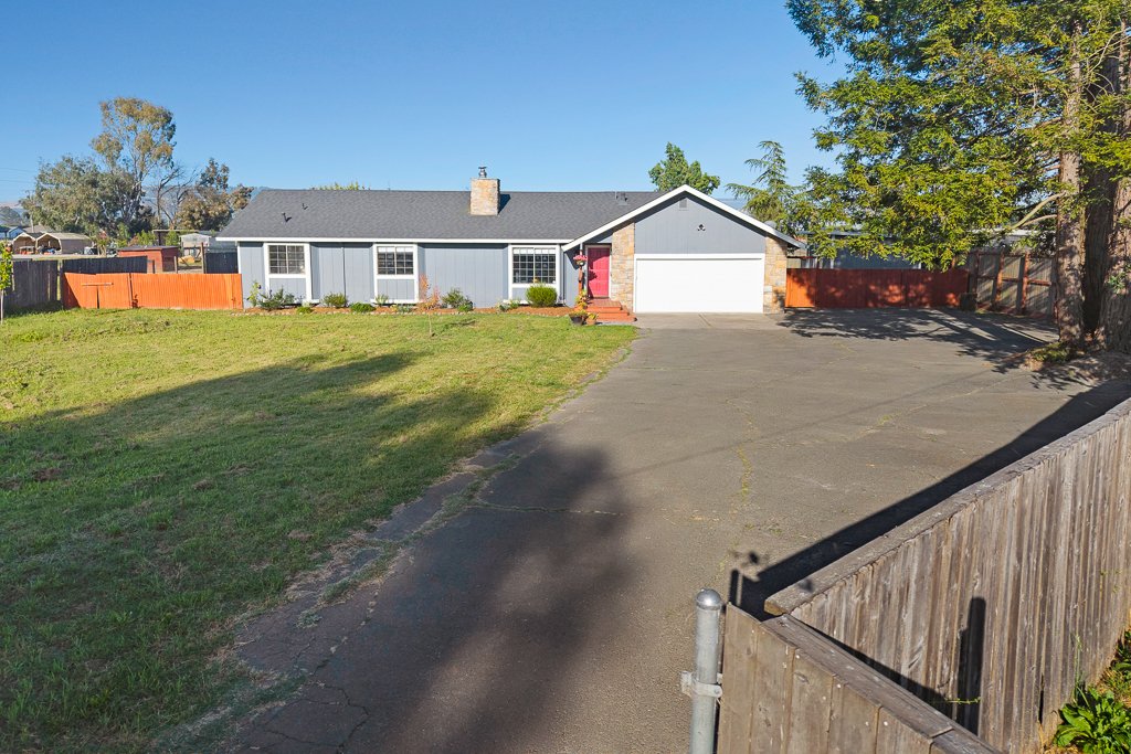 Front view of a suburban house with a driveway, green lawn, and some trees, under a clear blue sky.