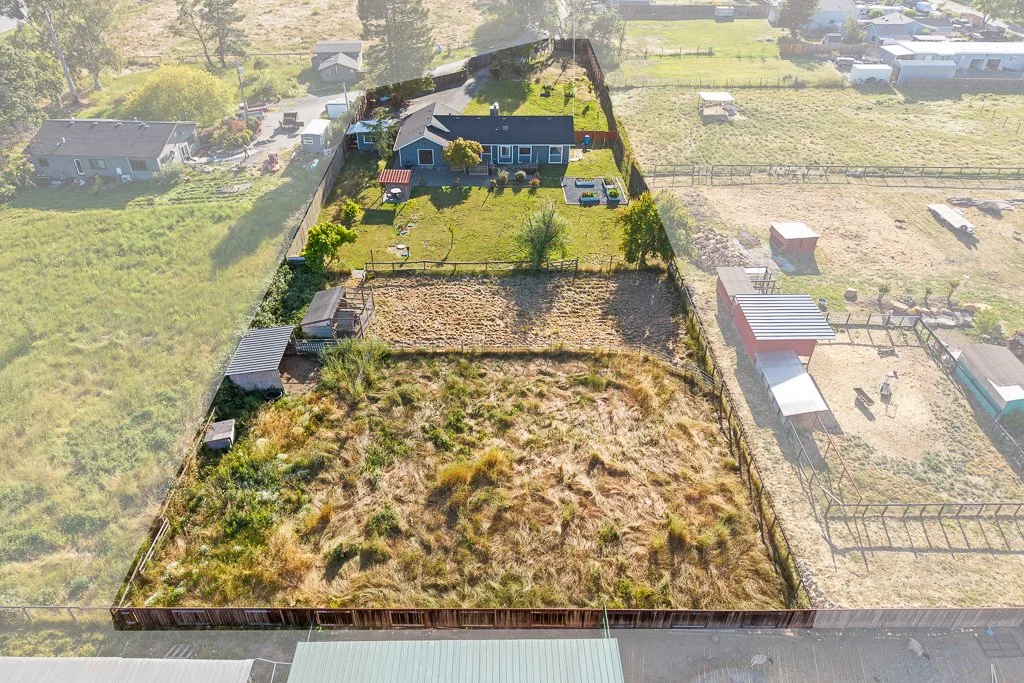 Aerial view of a backyard divided into sections with grass, garden plots, and fenced areas, adjacent to neighboring houses and open fields.