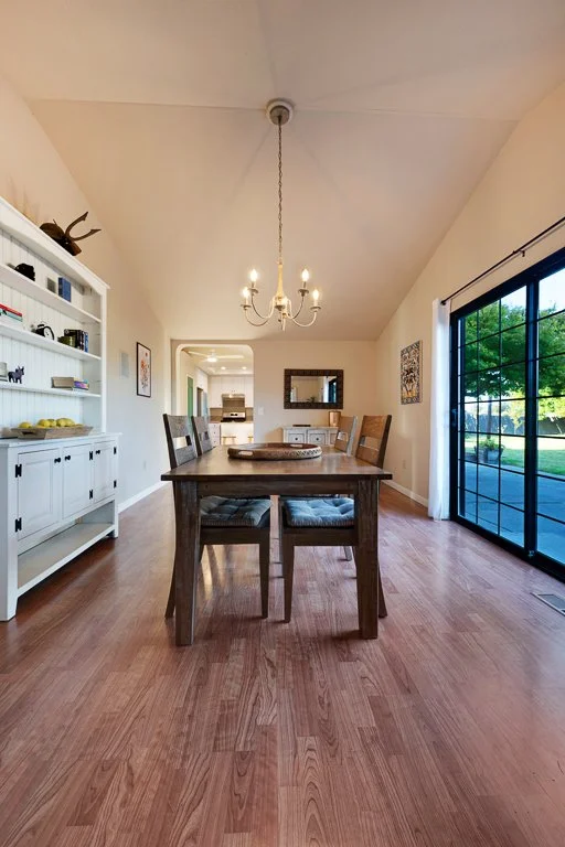 A dining room with a wooden table and four chairs, a white bookshelf on the left, a small cabinet at the back, a chandelier hanging above, a mirror and framed art on the wall, and sliding glass doors on the right showing a view outside.