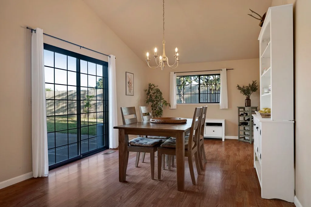 Dining room with wooden table, four matching chairs with upholstered seats, white curtains on sliding glass door and window, chandelier hanging from ceiling, white bookshelf with decor, small white cabinet, potted plant, and artwork on wall.