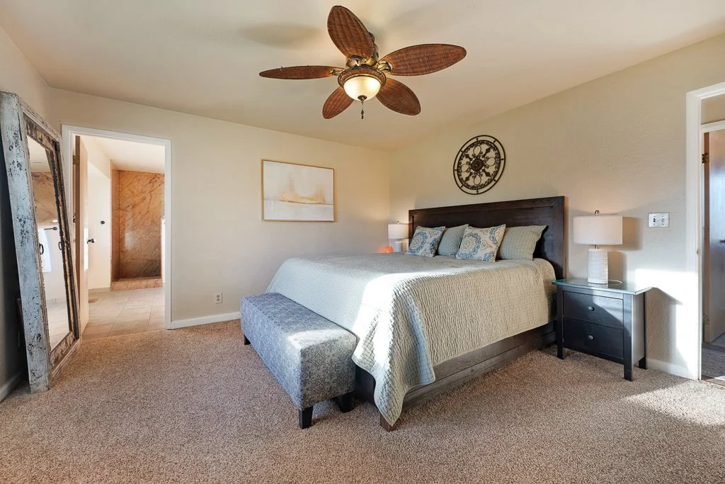 A bedroom with a king-sized bed, dark wooden headboard, beige bedding, patterned pillows, two side tables with lamps, a large mirror, artwork on the wall, a ceiling fan with leaf-shaped blades, and an open doorway leading to a bathroom.