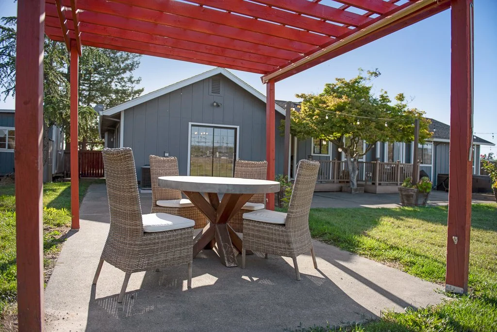 Outdoor patio area with a round table and four wicker chairs under a red pergola, behind a house with a shed, tree, and string lights.