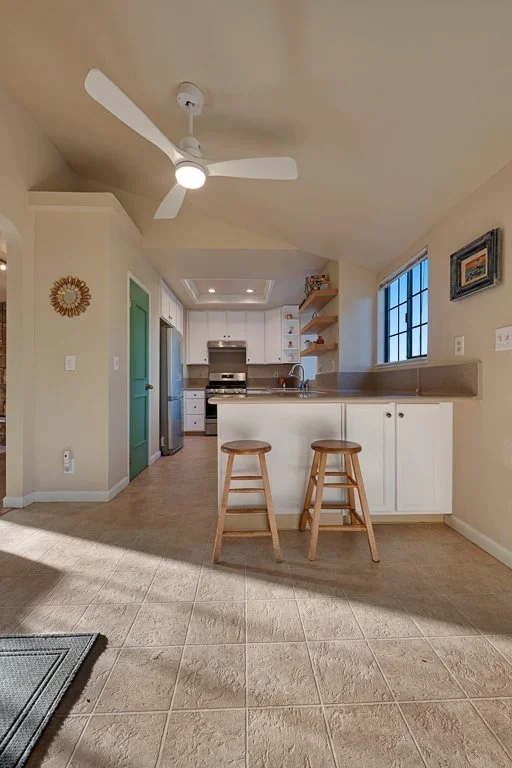 A bright, modern kitchen with white cabinets, a stainless steel refrigerator, and a stove. Two wooden stools are placed at a breakfast bar, and a ceiling fan is visible. There is a window on the right side and some wall decorations.
