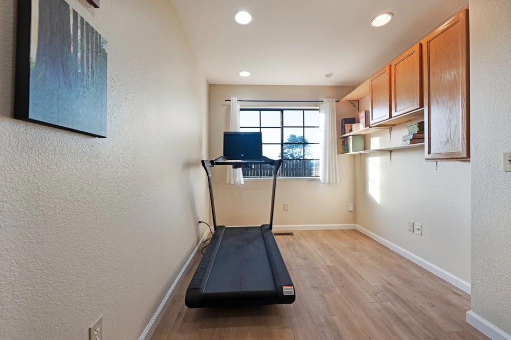 A small room with light-colored walls, wooden flooring, and a window with white curtains. There is a treadmill with a monitor in the center of the room, and built-in wooden cabinets and shelves with books on the right side.