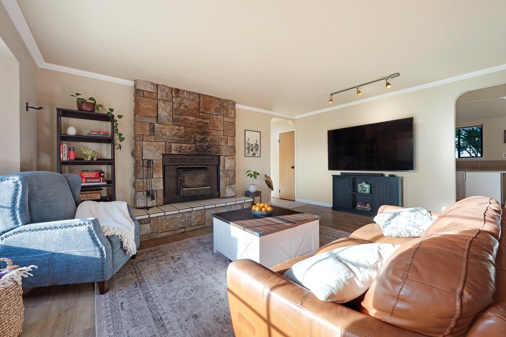 Cozy living room with a stone fireplace, blue armchair, tan leather sofa, coffee table, large flatscreen TV, and a bookshelf with decorative items and books.