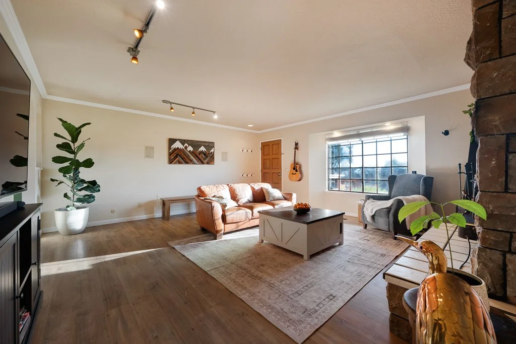 A spacious living room with hardwood floors, a beige sofa, a gray armchair, a white coffee table, a large window with blinds, a guitar hanging on the wall, a potted plant, and a stone fireplace on the right.
