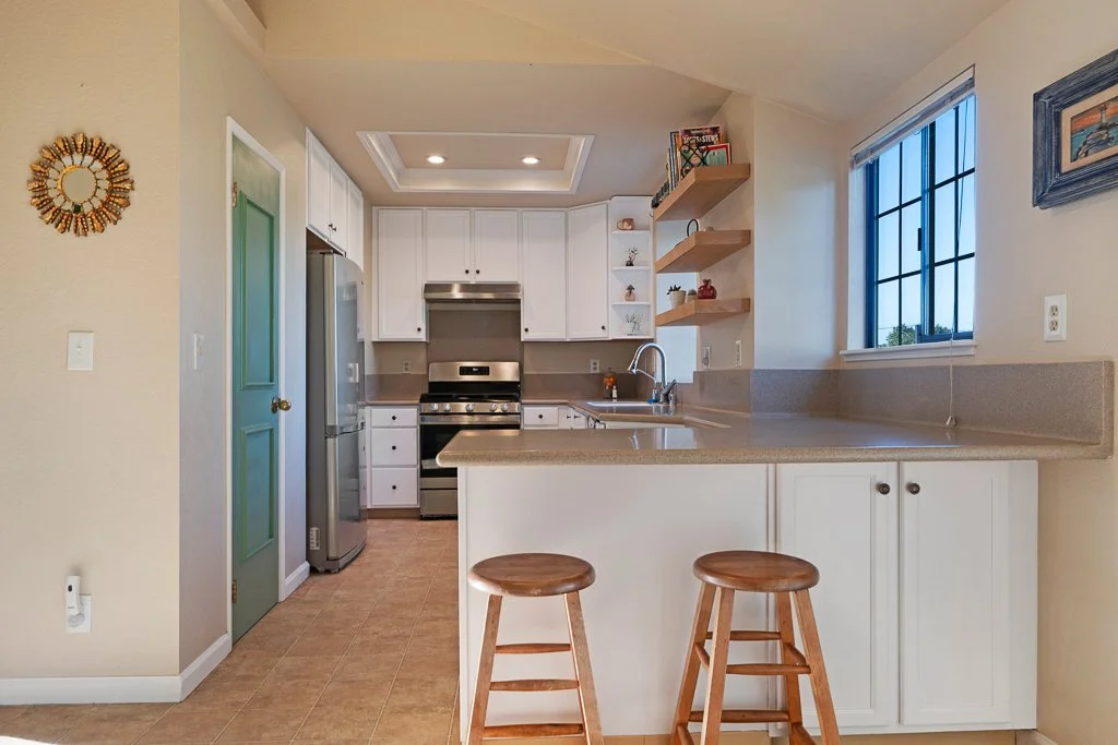 Bright kitchen with white cabinets, a stainless steel oven and refrigerator, open shelving, a window with black grid, and two wooden bar stools at a beige countertop.