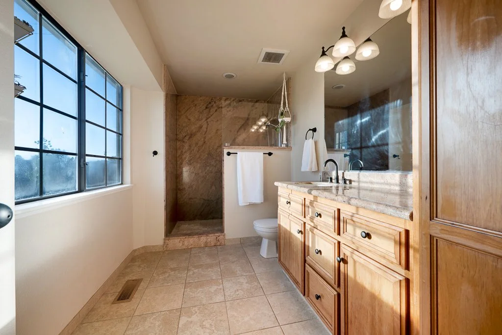Bathroom with large window, beige tile floor, wooden cabinets, granite countertop, double sink, mirror, towel rack, and shower area with marble walls.