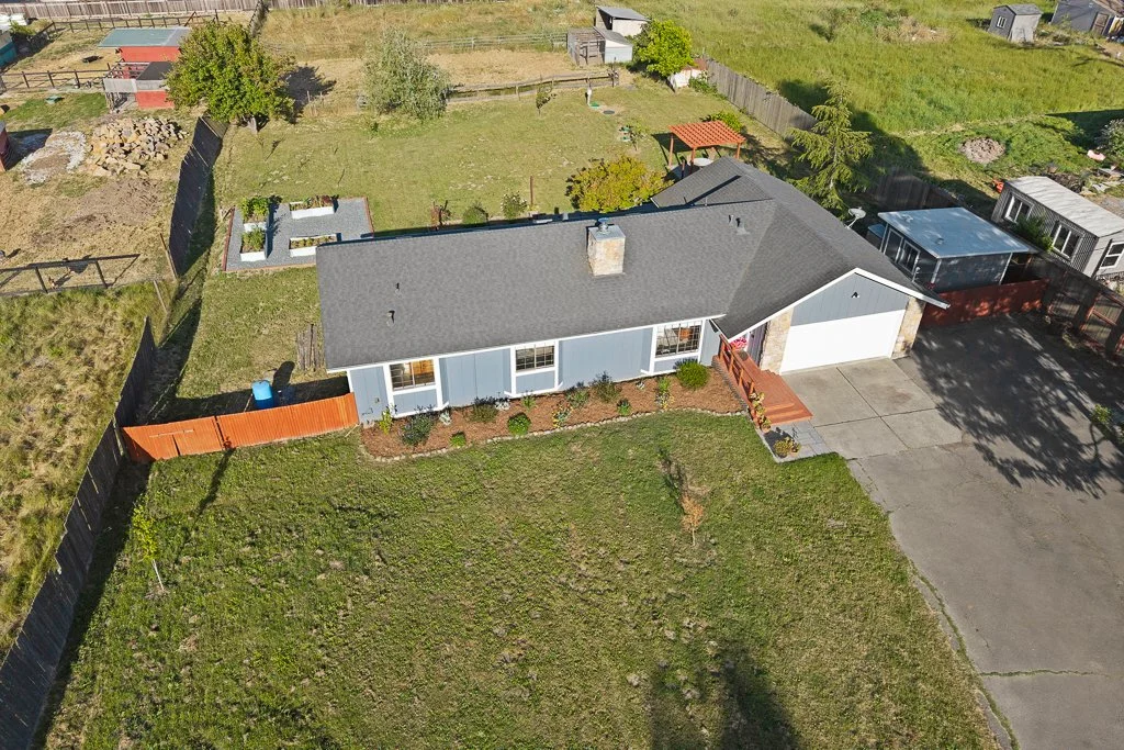 An aerial view of a single-story house with a gray roof, blue exterior walls, a driveway, and a small front porch with steps. The backyard has a garden, trees, and fenced sections.
