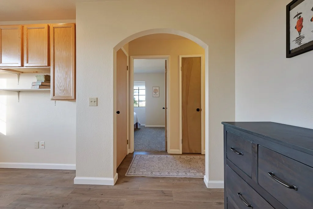 Interior view of a home with an arched doorway leading to a bedroom, kitchen cabinets on the left, and a dark gray dresser on the right. A framed picture hangs on the wall to the right.