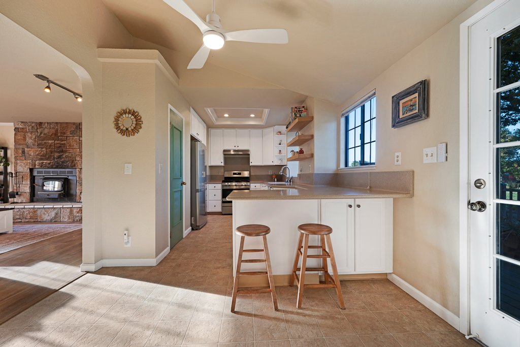 A bright kitchen with white cabinets, beige countertops, two wooden barstools, a ceiling fan, and large windows, with a view of a patio door leading outside.