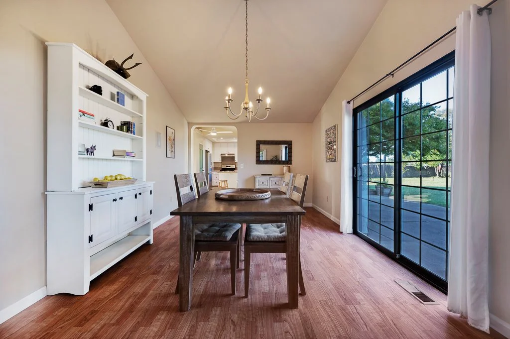 Dining room with wooden table and chairs, white bookshelf, chandelier, and large sliding glass door with curtains leading outside to a yard.
