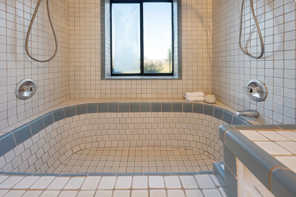 Tile-tub shower with two showerheads, a small window, and folded white towels on the ledge.