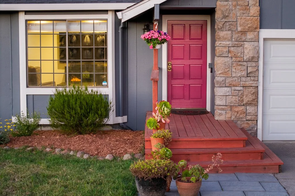 A front porch with a red wooden door, potted plants, and a hanging flower basket, with a window on the left and a garage door on the right.