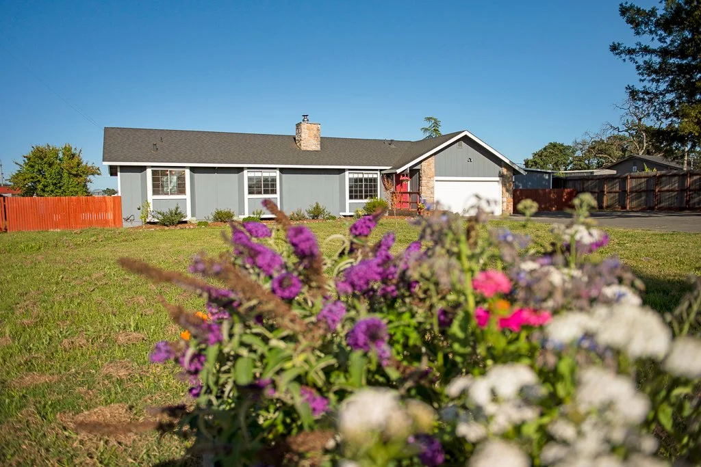 Front yard with flowers, a single-story house with grey siding, a brick chimney, and a parking area in the background under a clear blue sky.