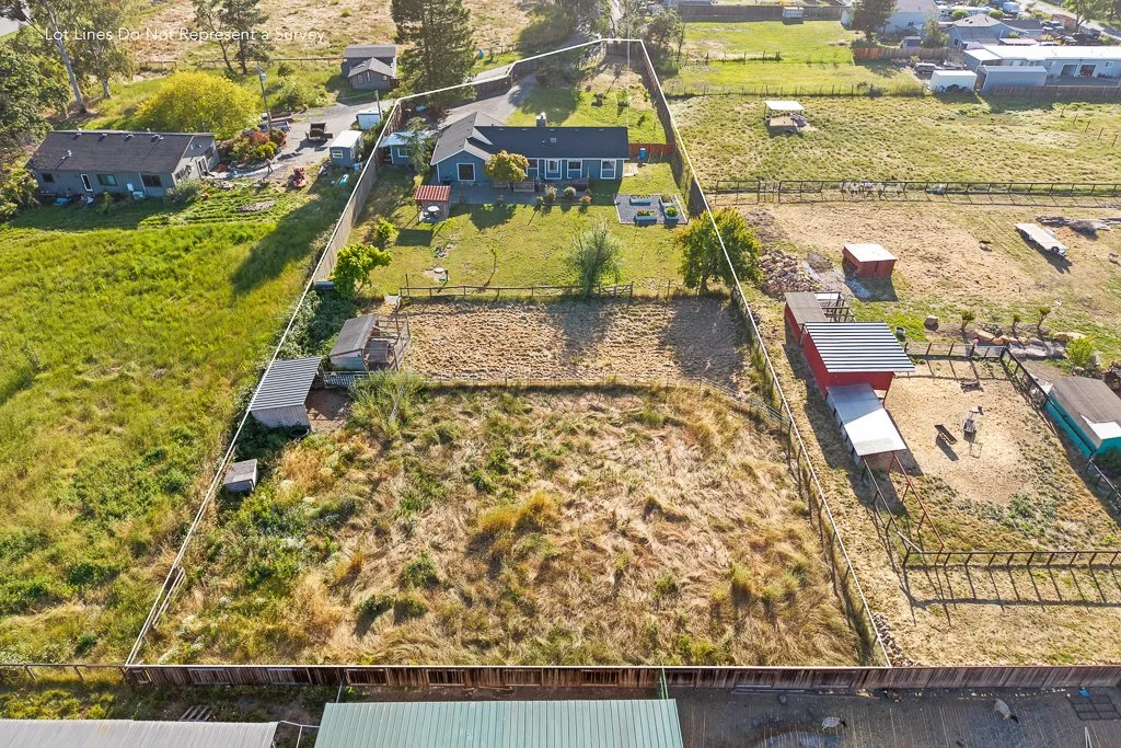 Aerial view of a backyard divided into sections with a house, garden, and fenced areas, surrounded by neighboring properties and open land.