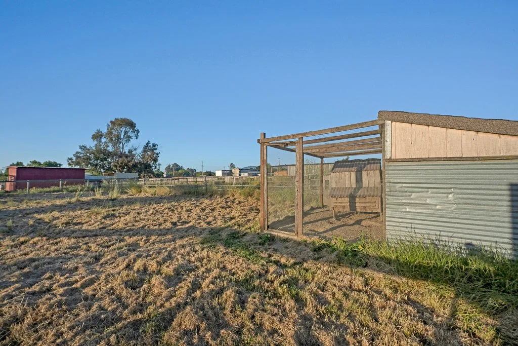 A small wooden and metal structure with a bench inside, situated outdoors on a dry, grassy plot of land under a clear blue sky.
