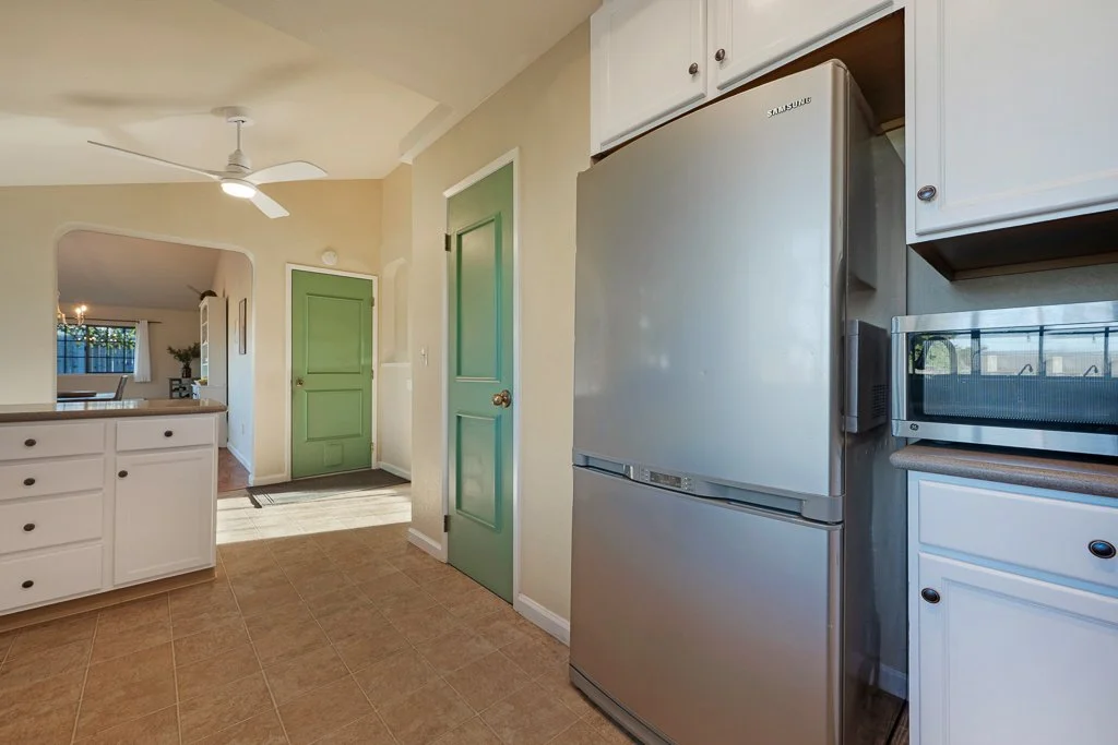 Kitchen with stainless steel refrigerator, white cabinets, microwave, green door, beige walls, and tiled floor, view from the kitchen looking toward the living area.