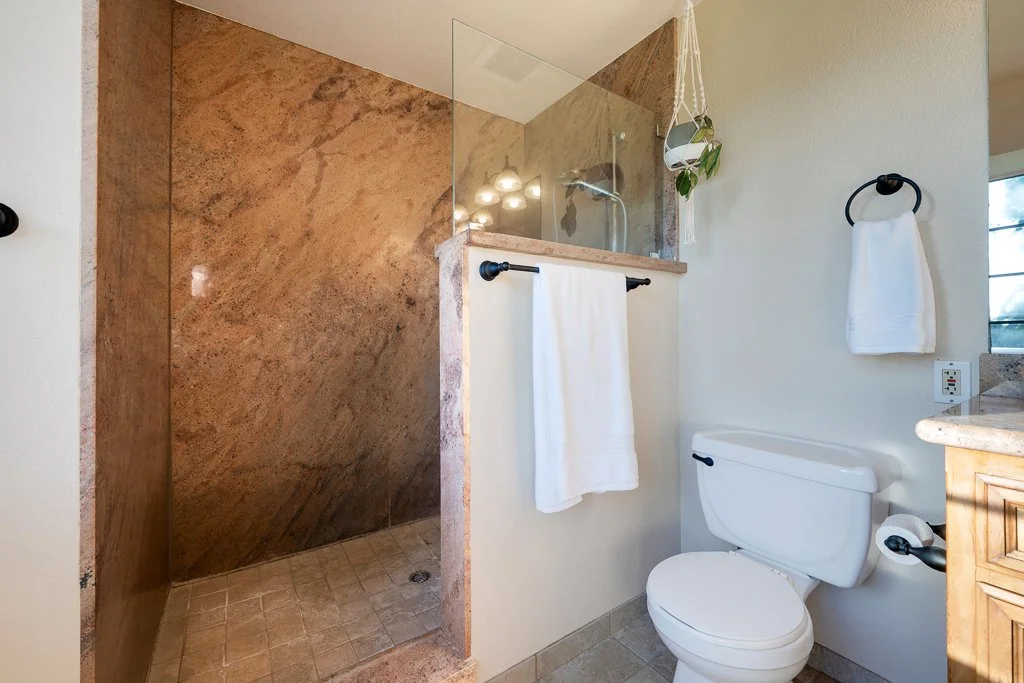 Bathroom with a walk-in shower that has a glass partition, a white toilet, and towels hanging on black towel rings. There is a small window with a view outside and a hanging plant near the shower.
