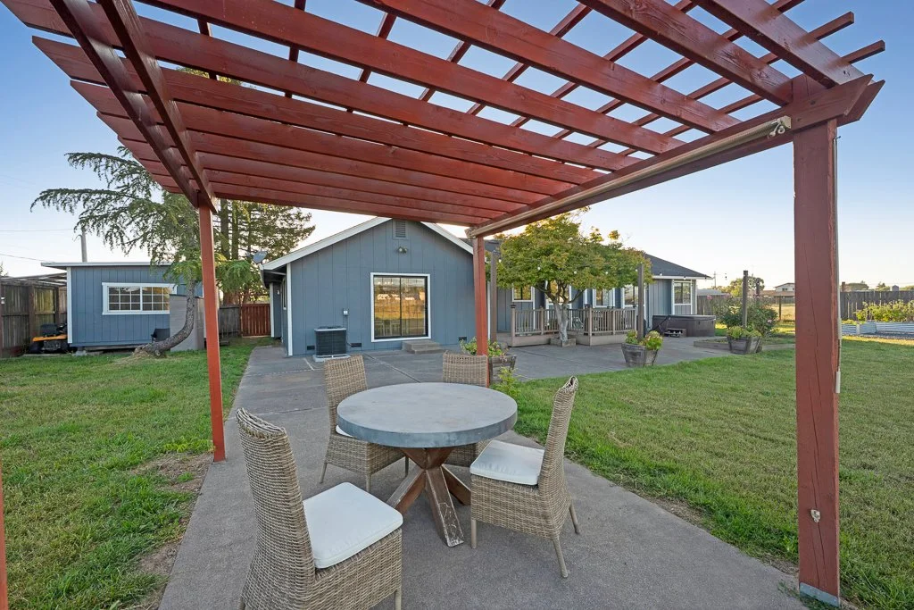 Backyard patio with a pergola, round table, four wicker chairs with white cushions, and a house with a deck, trees, and potted plants.