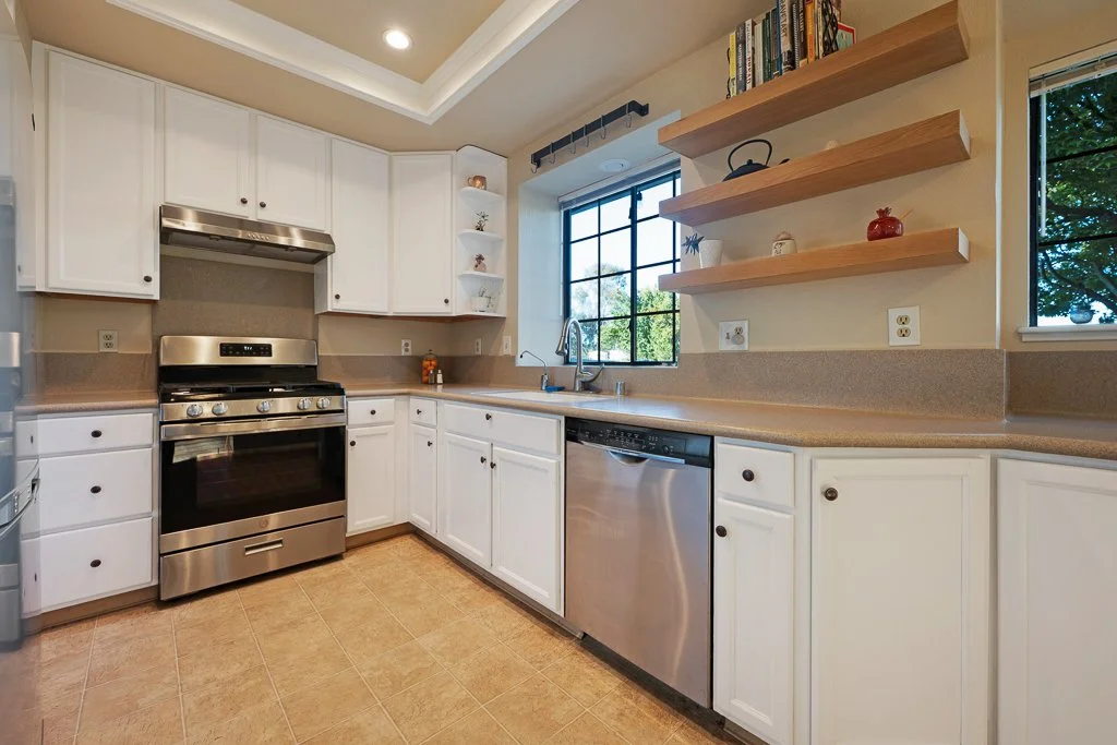 Modern kitchen with white cabinets, stainless steel oven and dishwasher, beige countertops, open wooden shelves, window with view of trees, and a yellowish tiled floor.