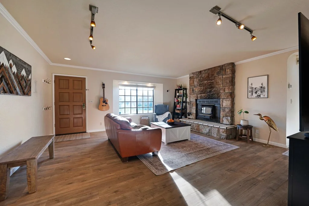 Living room with a wooden front door, guitar hanging on the wall, large window, brown sofa, blue armchair, fireplace with stone surround, bookshelf, artwork, and various decorative items.