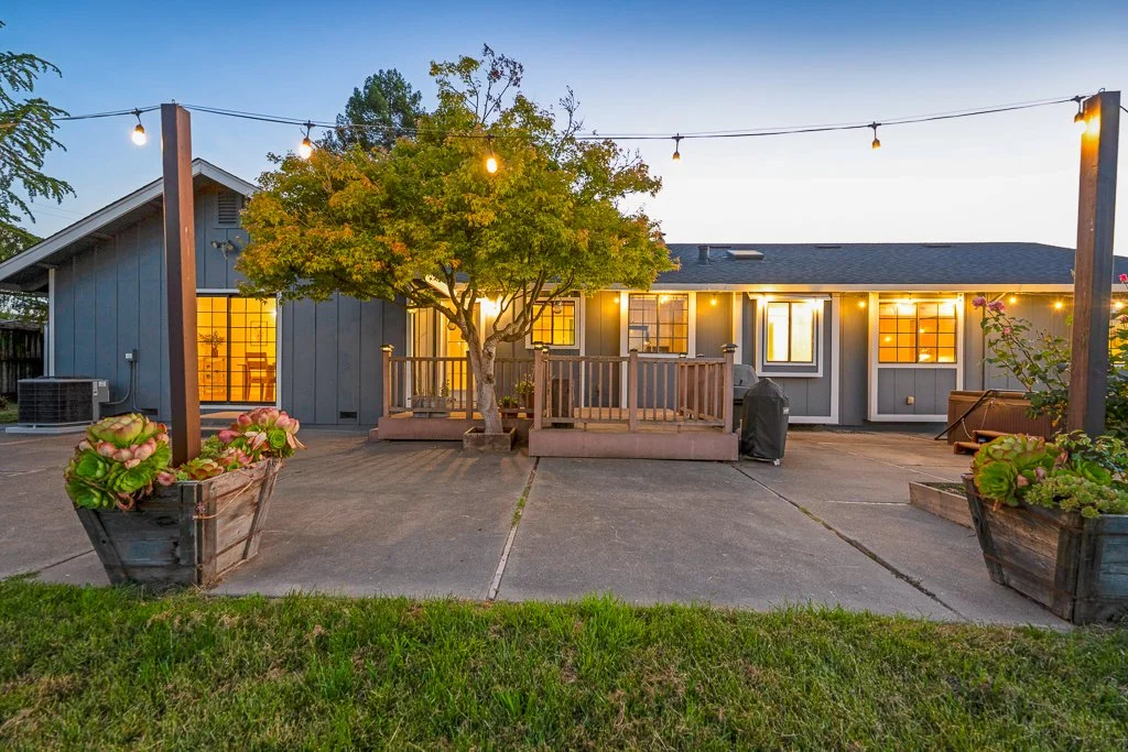 Backyard patio with string lights, potted plants, a central tree, a small deck, and a single-story house with lit windows.