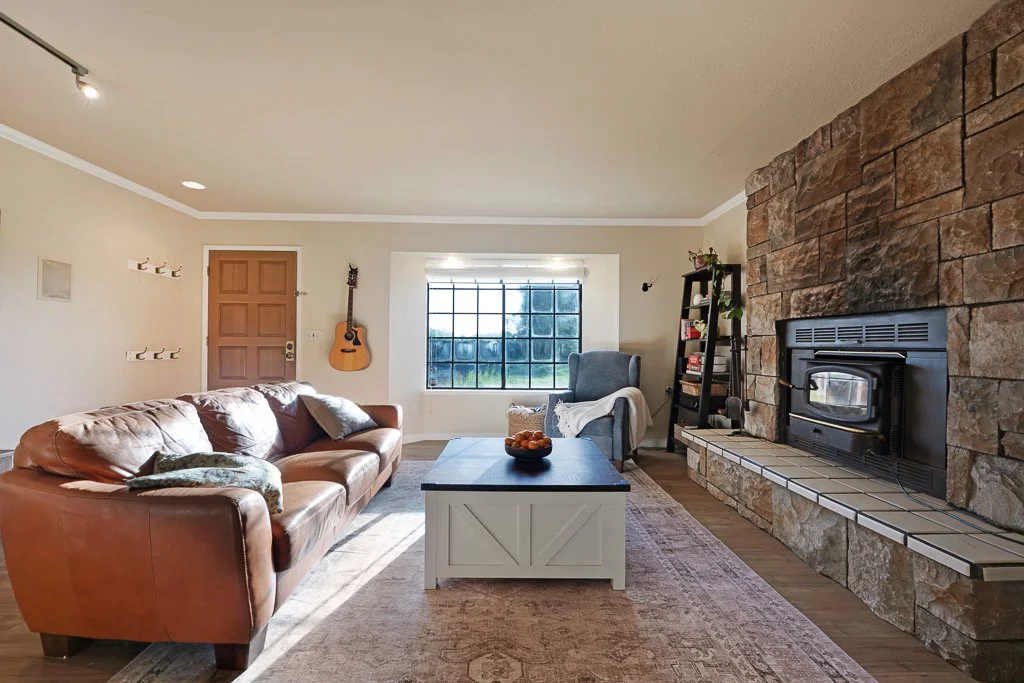 Living room with a leather sofa, gray armchair, stone fireplace, wooden coffee table, window, and guitar on the wall.