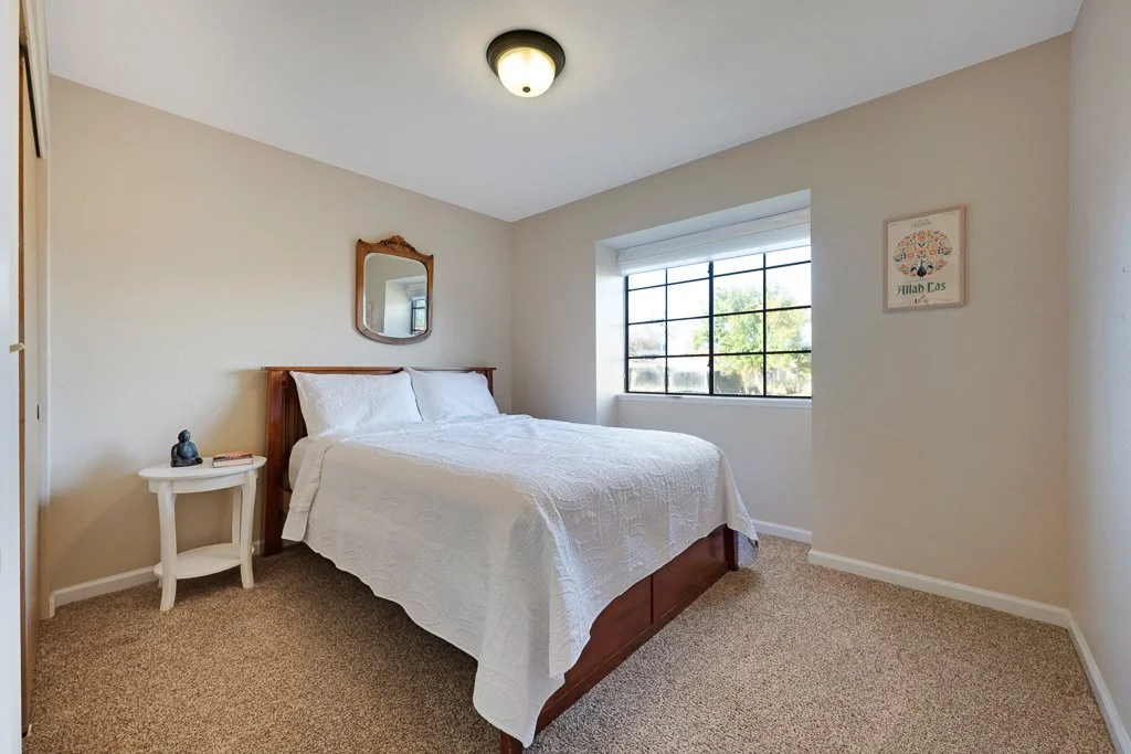 A minimalist bedroom with a wooden bed frame, white quilt, small white side table with a Buddha statue, and a large window with black frames letting in natural light.