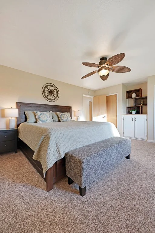 A bedroom with a large bed, dark wood headboard, beige bedding, and decorative pillows. There are two lamps on nightstands, a gray patterned bench at the foot of the bed, a wall clock, a ceiling fan with wooden blades, and a built-in cabinet with ope