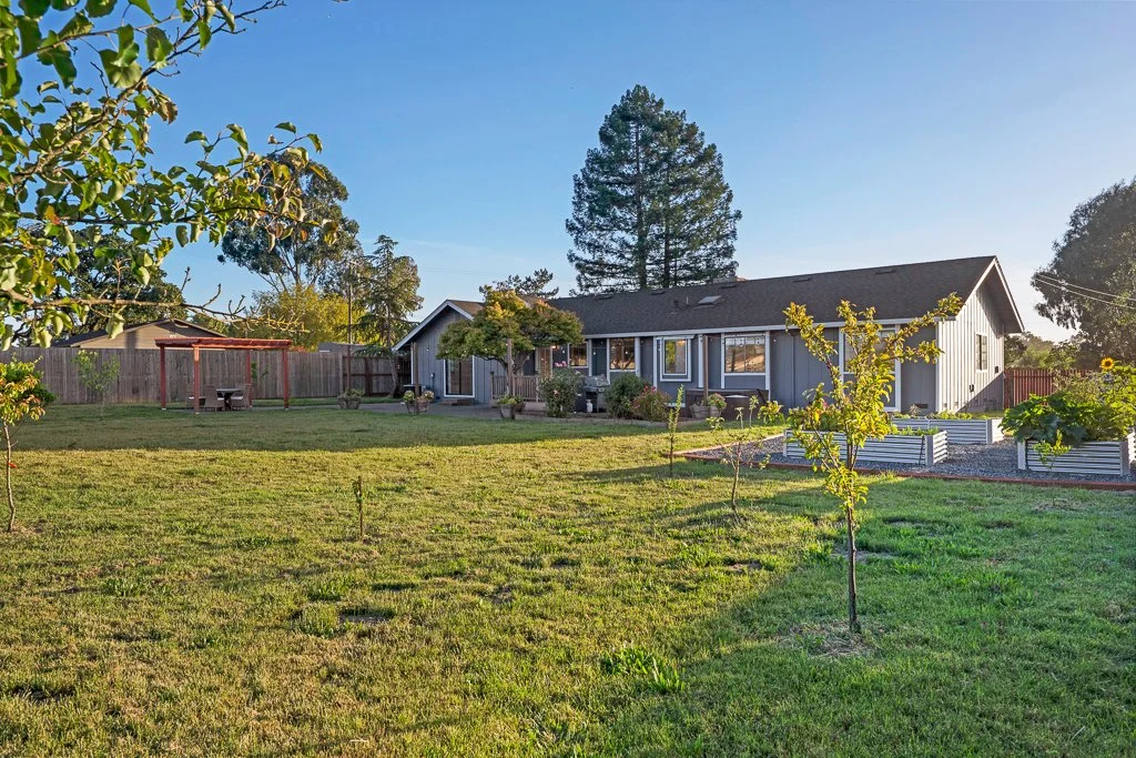 A backyard with a grassy lawn, small trees, a wooden swing set, and a gray house with multiple windows and a sloped roof, surrounded by a wooden fence and trees, under a clear blue sky.