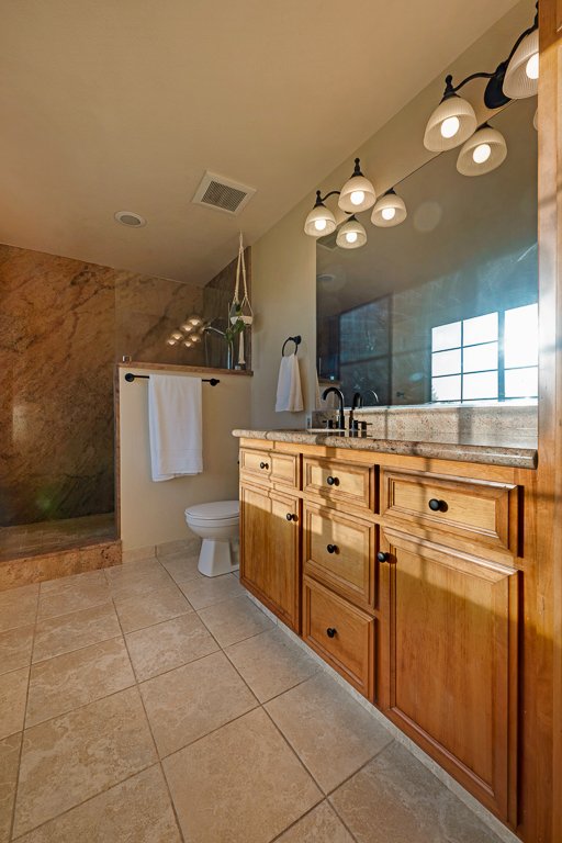 Bathroom with wooden vanity, granite countertop, large mirror, and modern lighting fixtures. Toilet next to the vanity and a shower area with a sliding glass door in the background.