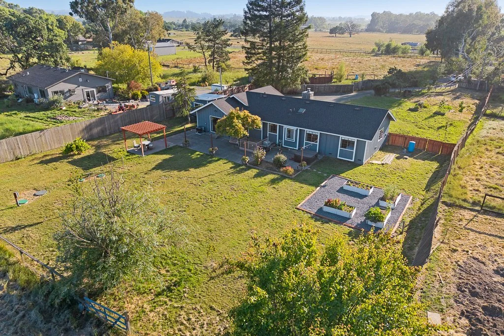 A spacious backyard with a well-maintained lawn, a small pergola with a table, a landscaped area with raised garden beds, and a blue house with a deck in a suburban neighborhood. Fences surround the yard with trees and open fields in the background.