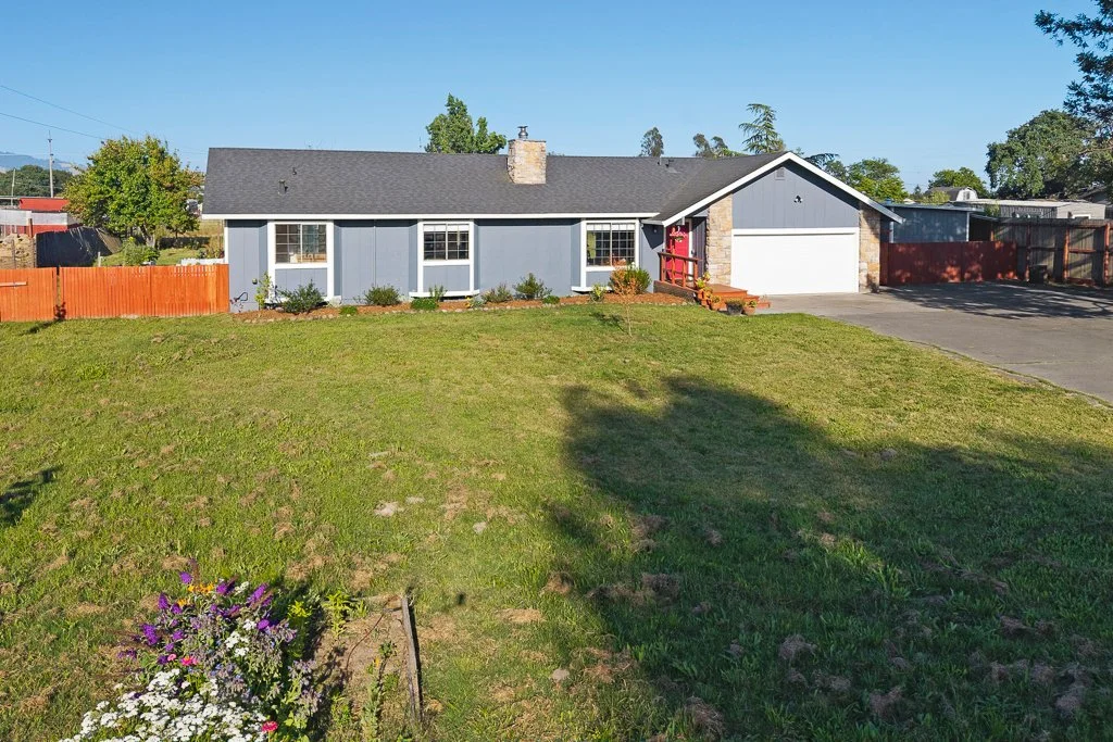 Front yard of a blue house with a gable roof, driveway, and red wooden fence, with trees and a clear blue sky in the background.