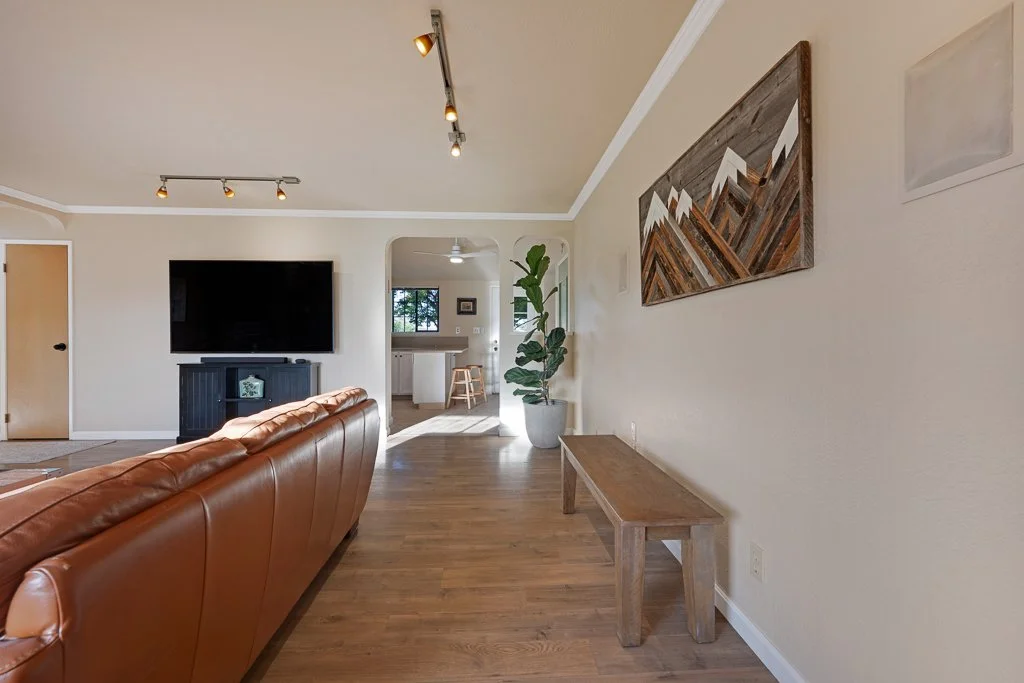 Living room with a leather sofa, a wall-mounted flat-screen TV, a wooden dining area visible through an archway, and a large potted plant, featuring minimalist decor.