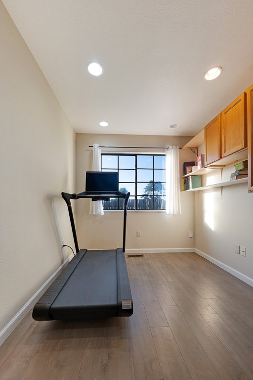 A room with a treadmill and a laptop on top, leading to a window with white curtains, wood flooring, and a wall-mounted shelf with books and wooden cabinets.