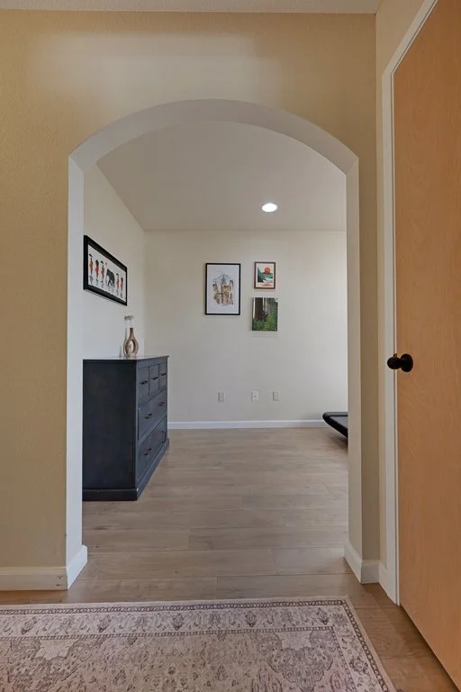 View through an arched doorway into a room with framed artwork on the wall, a black dresser with a decorative vase, and wooden flooring.