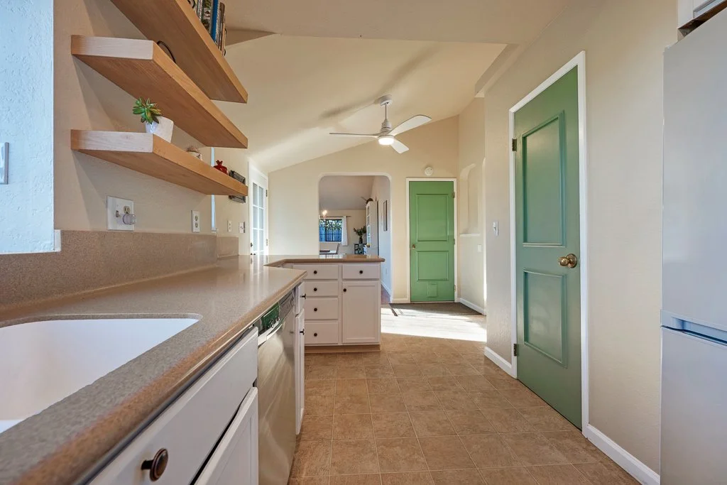 A bright kitchen with white cabinets, a beige countertop, and green doors. Open shelves on the wall hold small potted plants and decorative items. There is a ceiling fan and a view into another room with a window and sunlight.