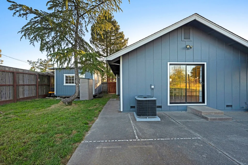 Backyard of a blue house with a concrete patio, a sliding glass door, a small set of stairs, an air conditioning unit, a tree leaning over the patio, and a wooden fence surrounding the yard.