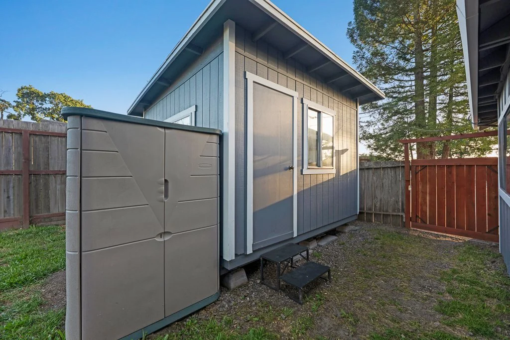 Backyard shed with gray exterior, small steps leading to door, wooden fence, large plastic storage container, and trees in the background.