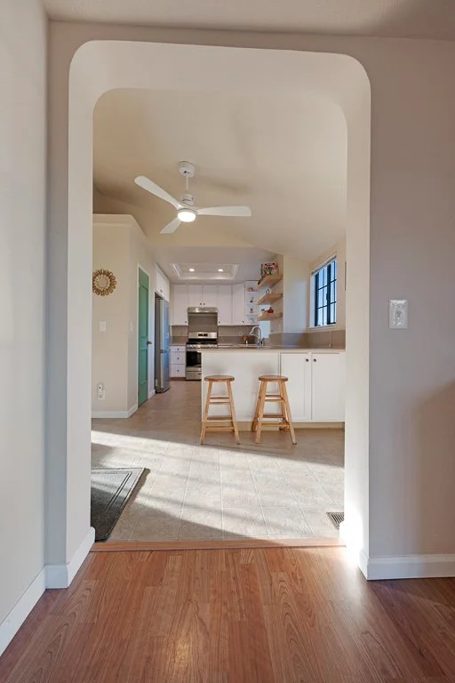 View of a kitchen through an arched doorway, featuring a white ceiling fan, white cabinets, a window, and two wooden bar stools at the counter.
