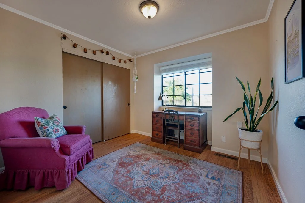 A cozy room with a pink armchair, a decorative pillow, a wooden desk near a large window, a potted plant, and a patterned area rug on hardwood floors.