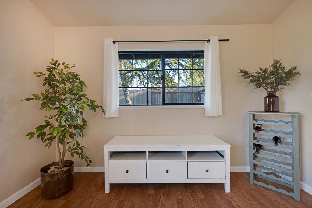 A small room with a window, white curtains, a potted plant on the left, a white storage bench in the center, and a wine rack with a plant on top on the right, with hardwood flooring.