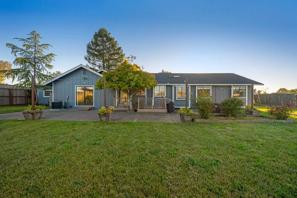 Rear view of a single-story gray house with a wooden deck, surrounded by a green lawn and potted plants, under a clear sky.