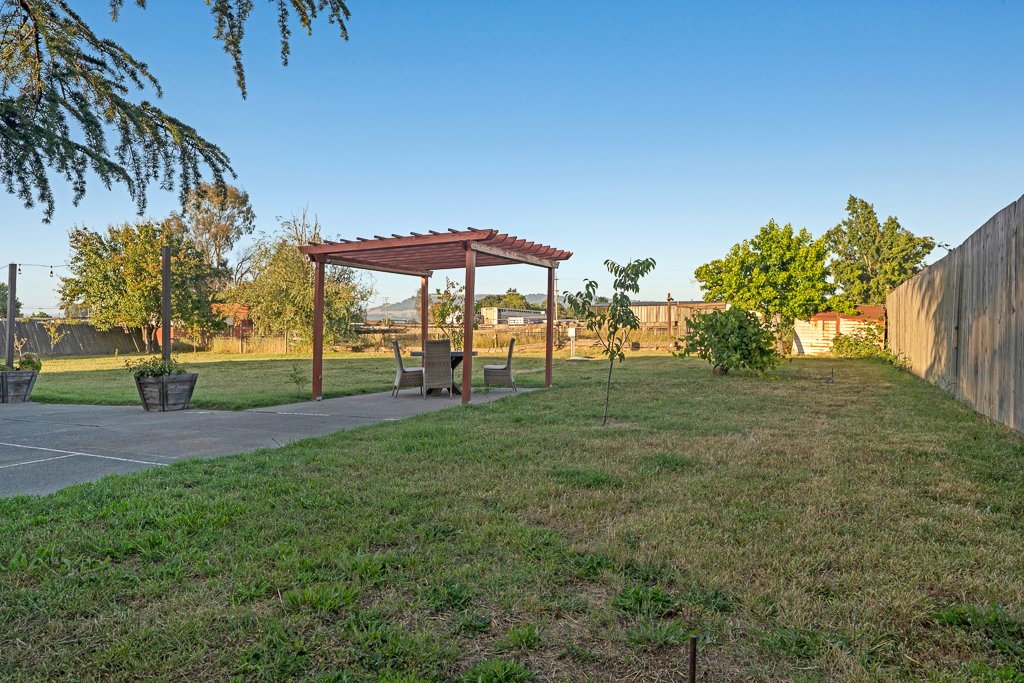 A backyard with a grassy lawn, small trees, a concrete patio, and a wooden pergola with outdoor furniture underneath. There are wooden planters, a wooden fence on the right, and a clear blue sky.