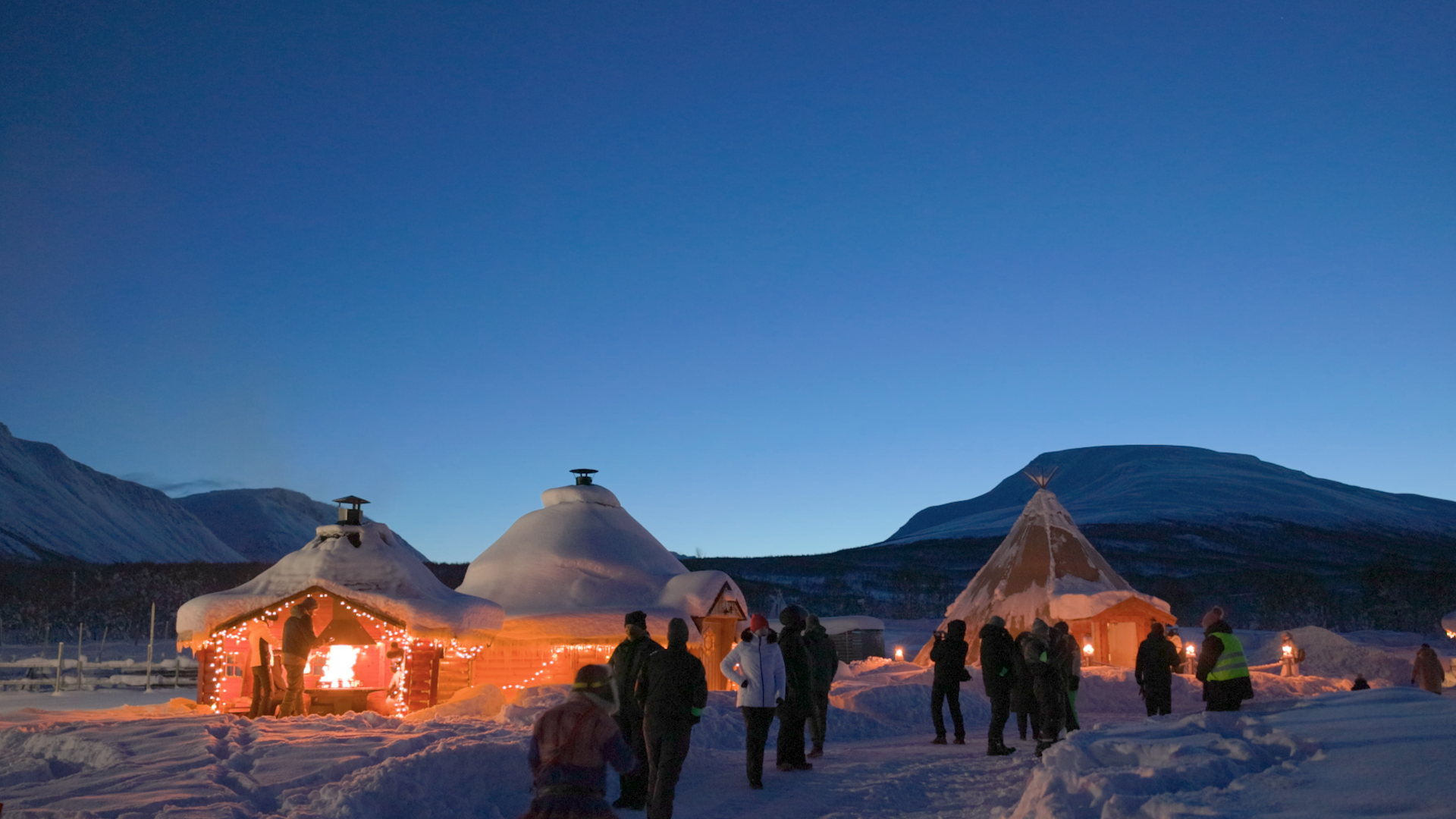 Snow-covered open-air structures with fire pits inside, tourists visiting a Sami reindeer herding community